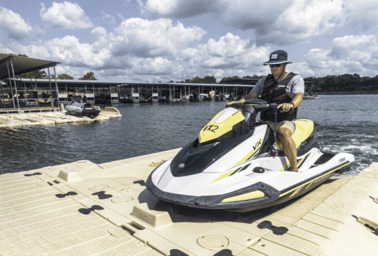 A person wearing sunglasses and a bucket hat rides a yellow and white jet ski onto a floating dock at a marina, with other watercraft and covered docks visible in the background under a partly cloudy sky.