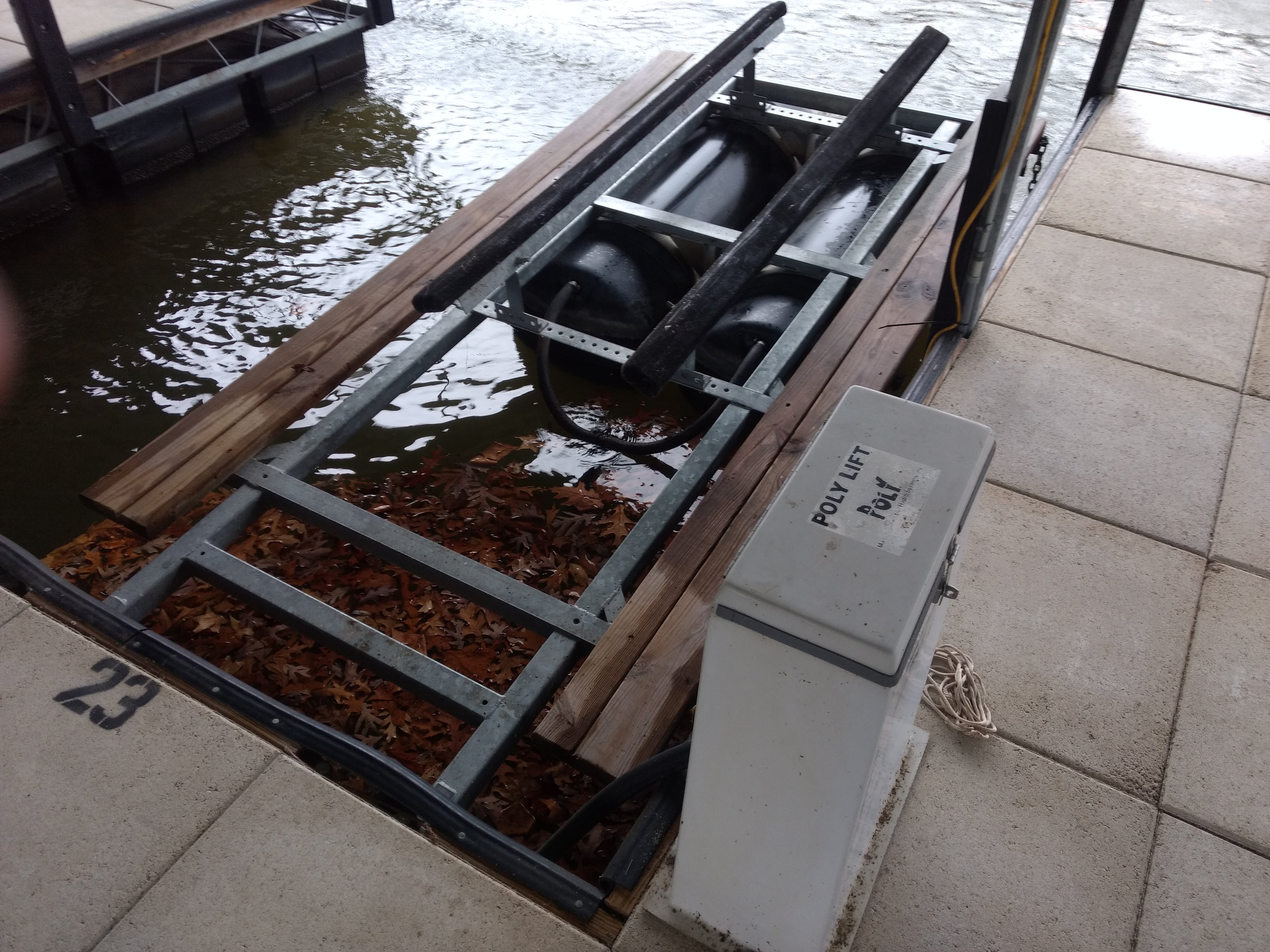 Poly Lift® PWC Front Lift by Poly Lift shown over water at a dock features a metal frame, black pontoons, wooden planks, and fallen leaves below. A control box marked POLY LIFT ROLL is mounted on the concrete dock nearby.