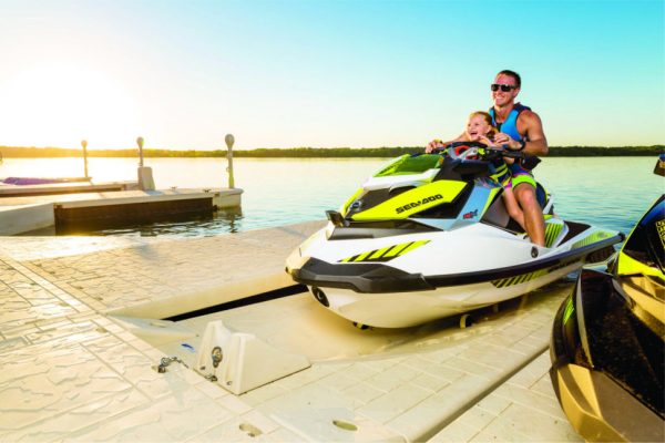 A man and a young child smile while sitting on a yellow and white Sea-Doo jet ski at the dock, enjoying the easy-to-use Poly Lift Wave Armor SLX5 port with its independent wheel system. Calm water and a clear sky complete the sunny scene.