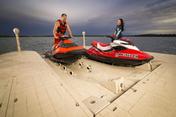 A man and a woman, both smiling, stand on jet skis parked on a beige Poly Lift Wave Armor SLX5 floating dock beside a large body of water under a cloudy sky.
