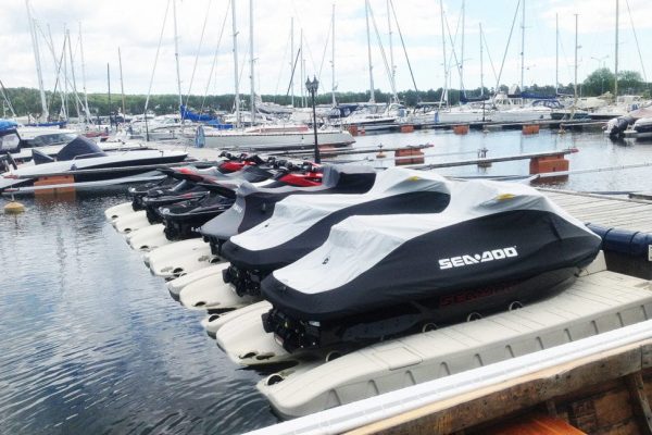 Several covered Sea-Doo jet skis are lined up on Poly Lift Wave Armor SLX5 floating docks at a marina, with sailboats and yachts moored in the background under a partly cloudy sky.